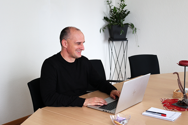 Ein lächelnder Mann sitzt an einem großen Konferenztisch und arbeitet auf einem Laptop; neben ihm liegen ein Notizblock und Stifte, im Hintergrund steht eine Pflanze auf einem hohen Ständer.